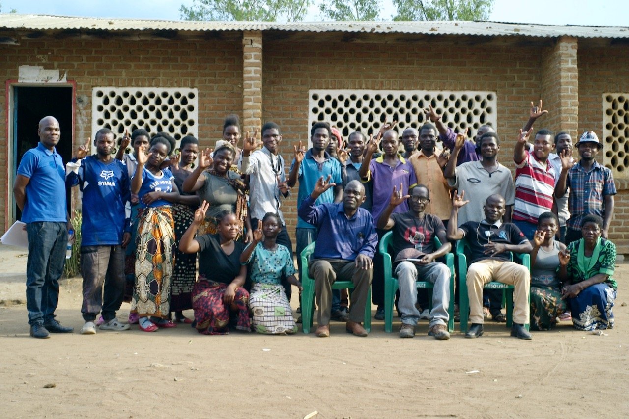 Iso joukko malawilaisia poseeraa ulkona tiilirakennuksen edessä. Suurin osa seisoo, edessä kolme miestä istuu vihreillä muovituoleilla ja neljä naista on kyykyssä. Suurin osa on nostanut kätensä tervehdykseen.